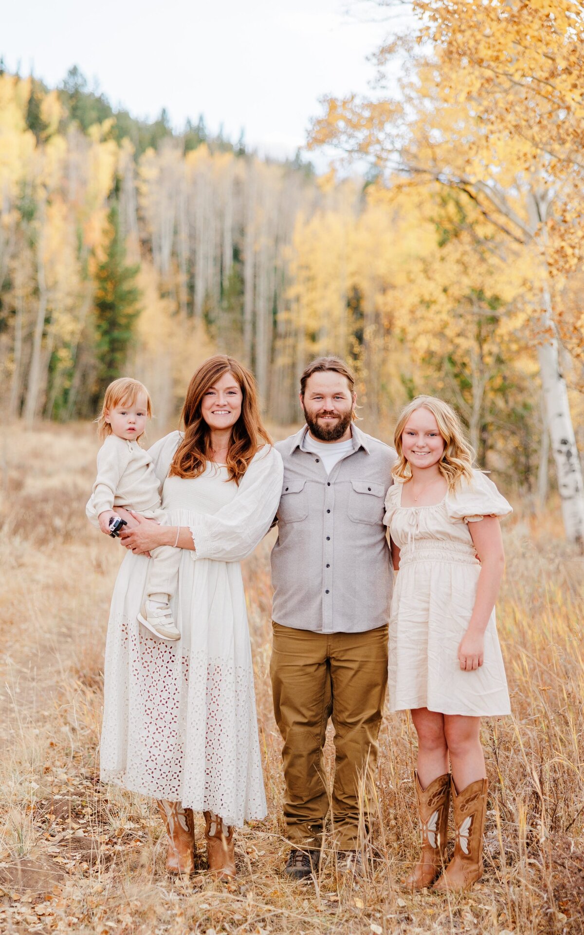 A happy mom and dad stand in a forest in fall with their teen daughter and baby on mom's hip