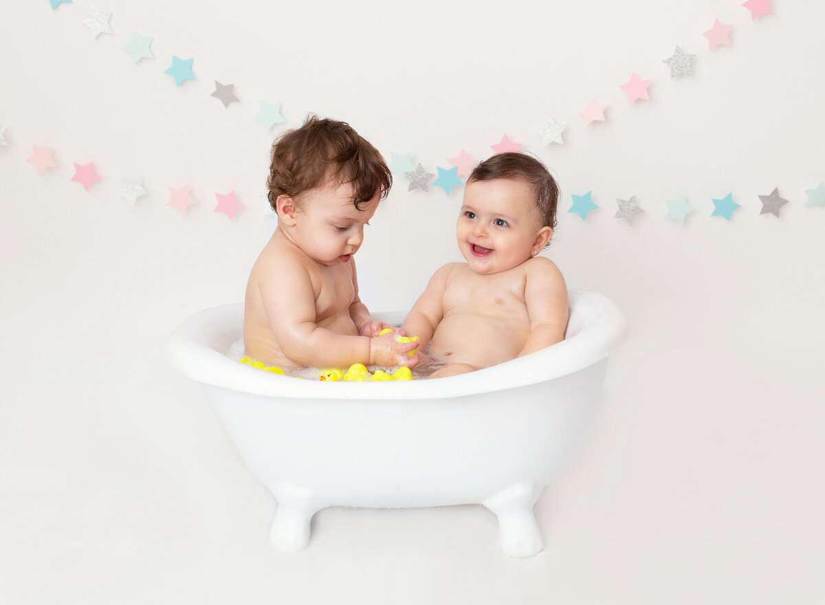 Baby boy and baby girl sit in a mini bathtub to clean up from a first birthday cake smash photoshoot. Babies are laughing and playing with rubber ducks.