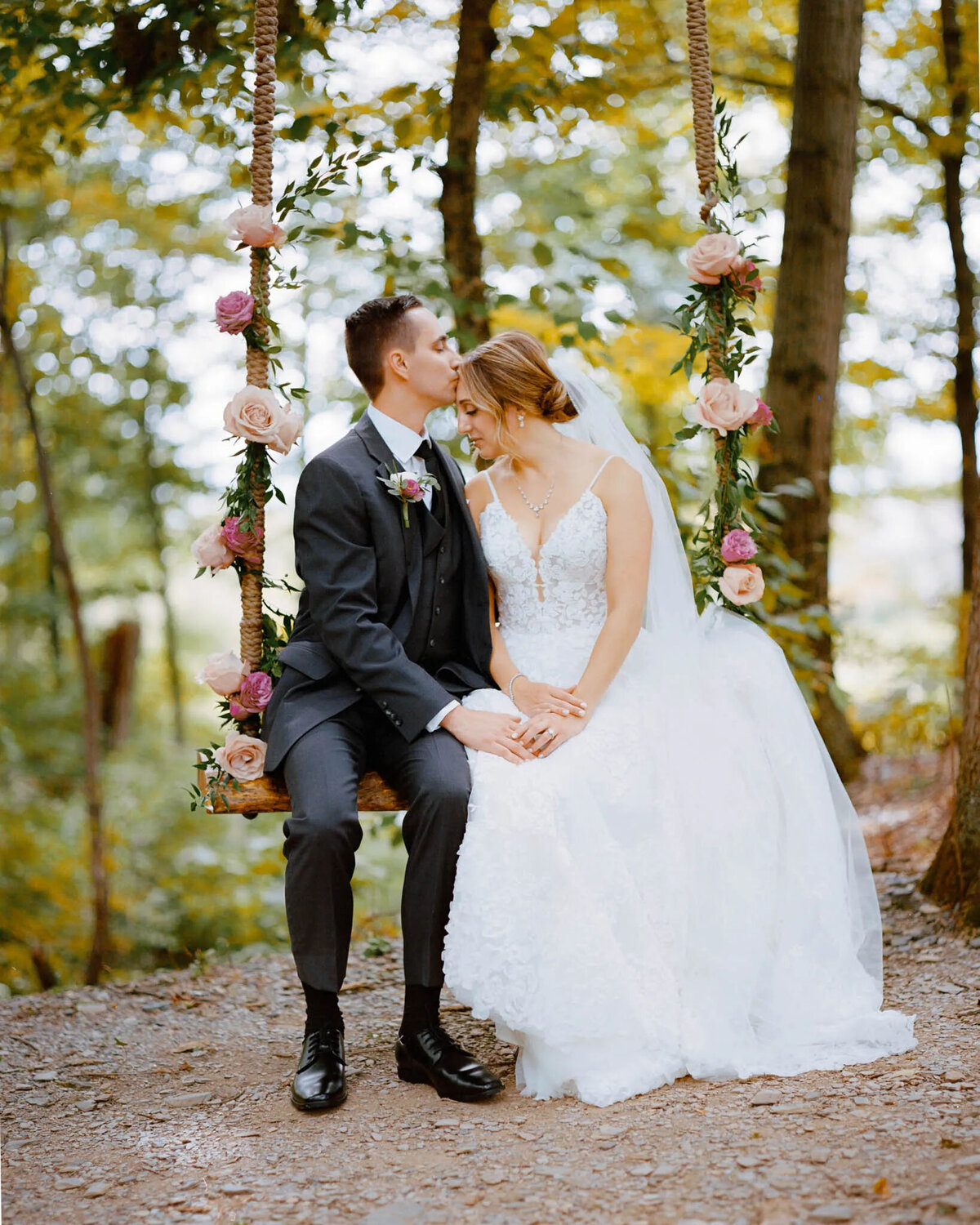 A bride and groom sit together on a flower-decorated swing in a forest. Captured by an NJ wedding photographer, the groom kisses the bride’s forehead while holding her hand. Both are dressed in formal wedding attire and appear calm and happy.