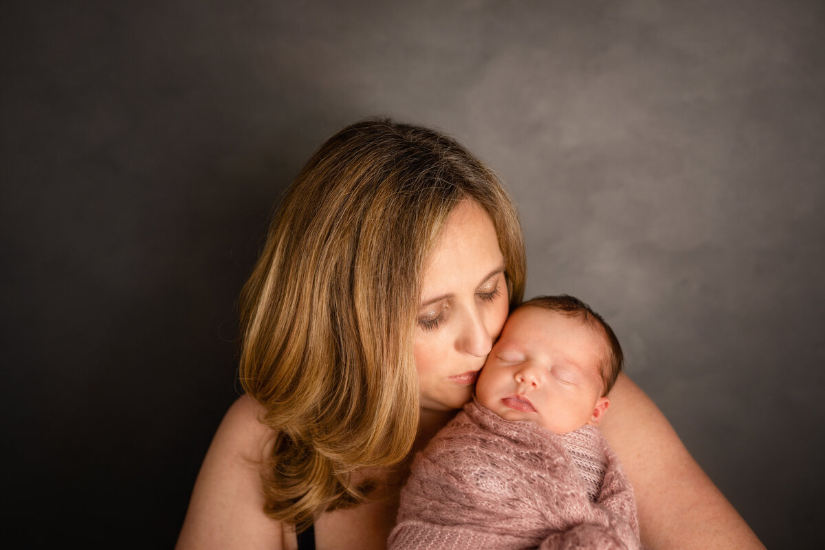 Mom with shoulder length blonde hair is kissing newborn baby girl on her cheek.  Mom is looking at baby and holding her in her arms.  Baby is wrapped in a pink swaddle.