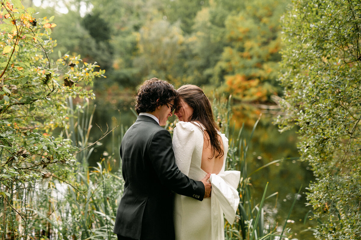 Bride and groom smiling with heads leaned in and lake in the back