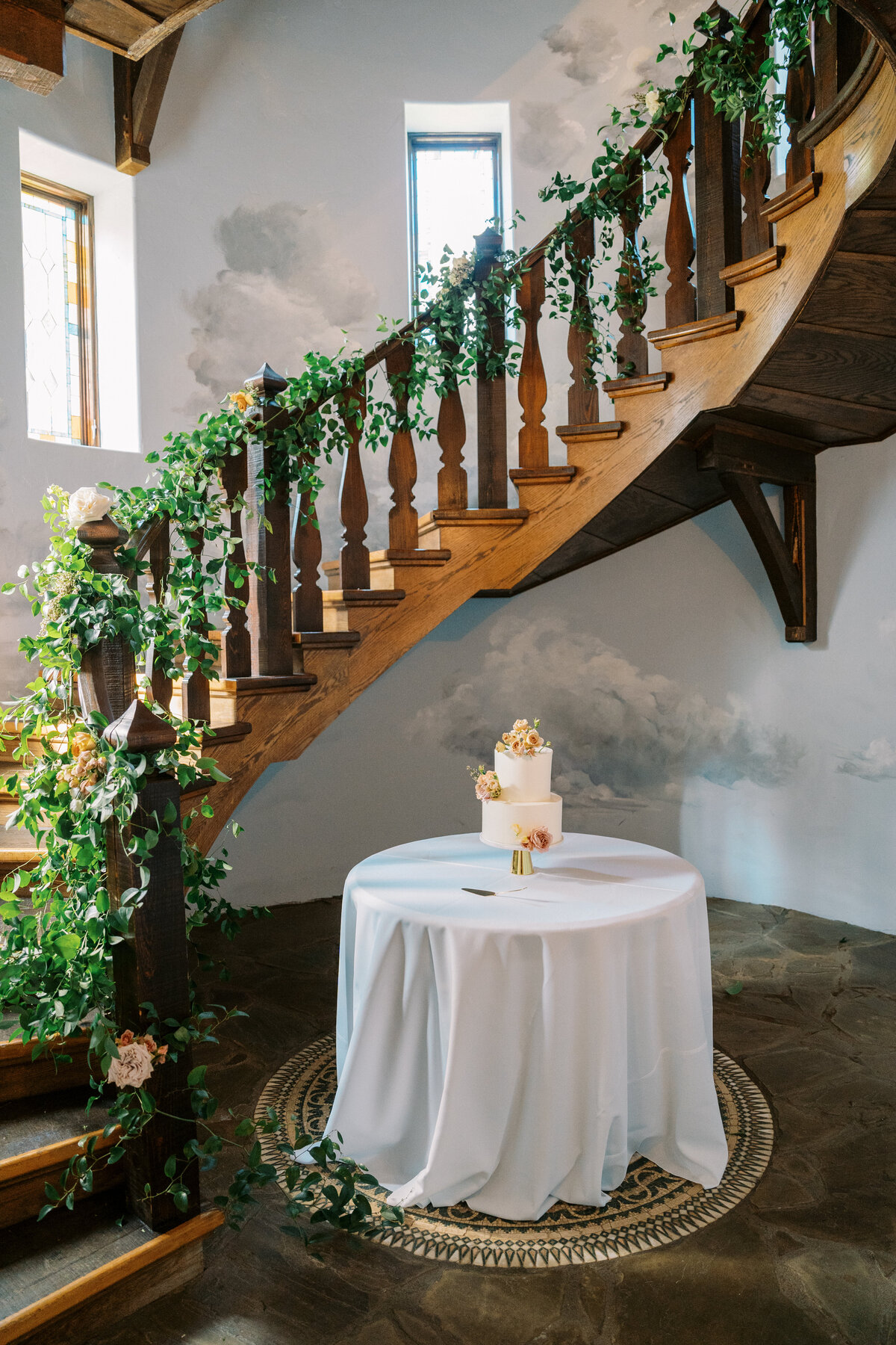 Wedding cake display placed at the base of a grand staircase wrapped in greenery and florals at Castle Ladyhawke.