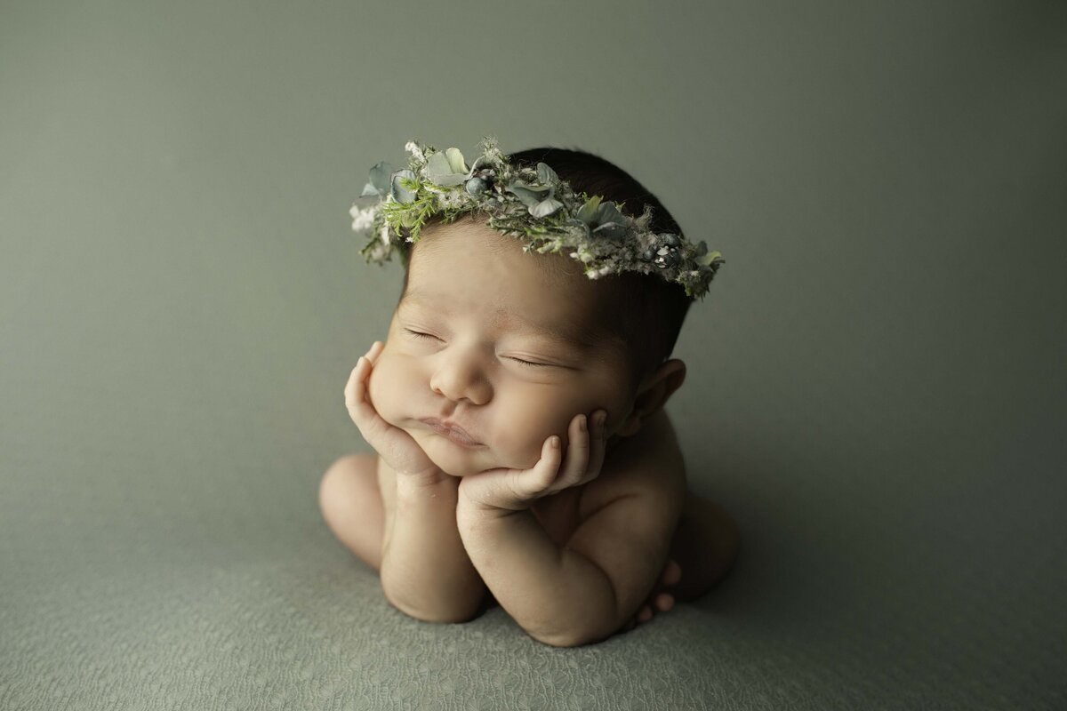 Newborn baby girl posed in the classic froggy position, her tiny hands resting under her chin, wearing a delicate blue flower halo. She peacefully rests on a soft backdrop, creating a dreamy and elegant portrait