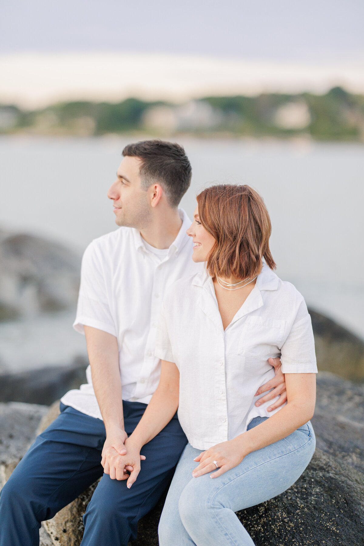 A close up of an engaged couple sitting on rocks at Wingaersheek Beach during sunset.