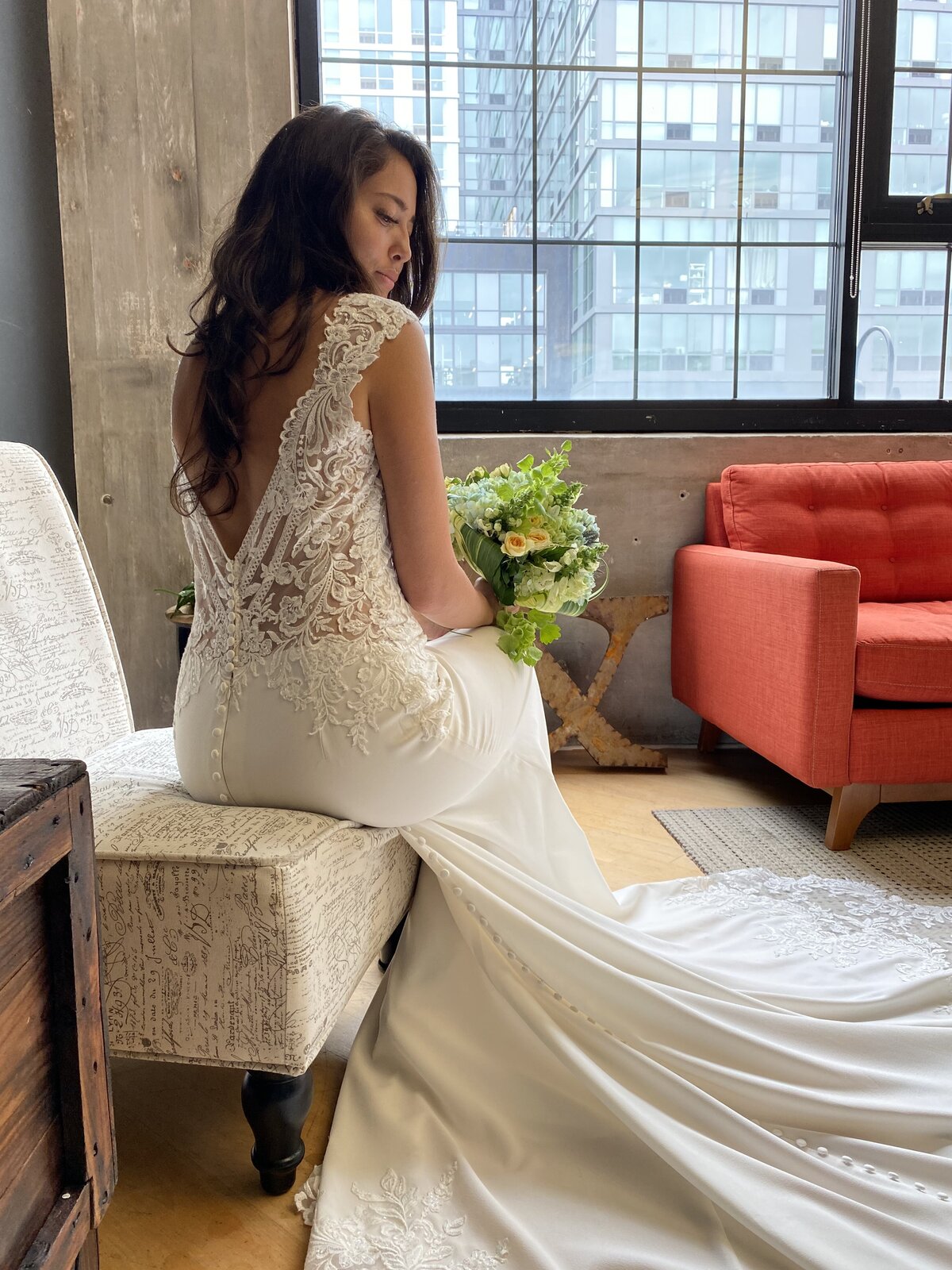 Bride stands near a window, showing off her long train and deep V-back wedding dress.