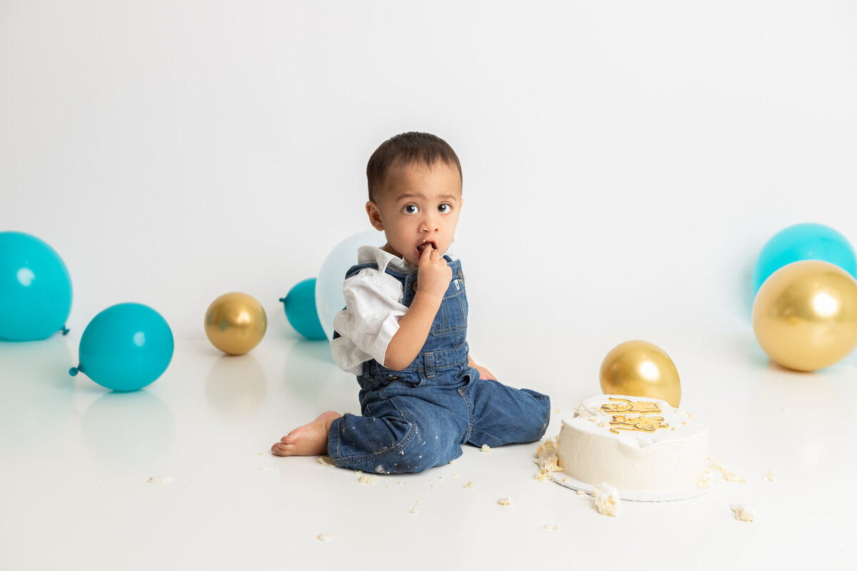 Studio cake smash for a boy with a white back drop and blue teal and gold balloons