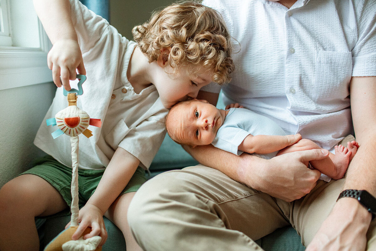 Close-up of brother kissing baby with family nearby indoors - Sara James San Antonio newborn photographer captures sibling love and new family dynamics during tender lifestyle sessions in comfortable home environments