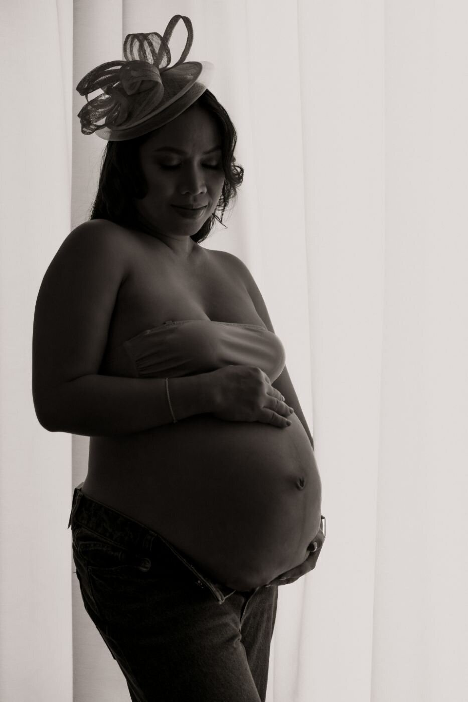 A close-up, color photo of an expecting mother looking down at her baby bump with a warm smile. She is wearing a white or light-colored shirt, and her hands are gently cradling her belly. Her expression is loving and serene.