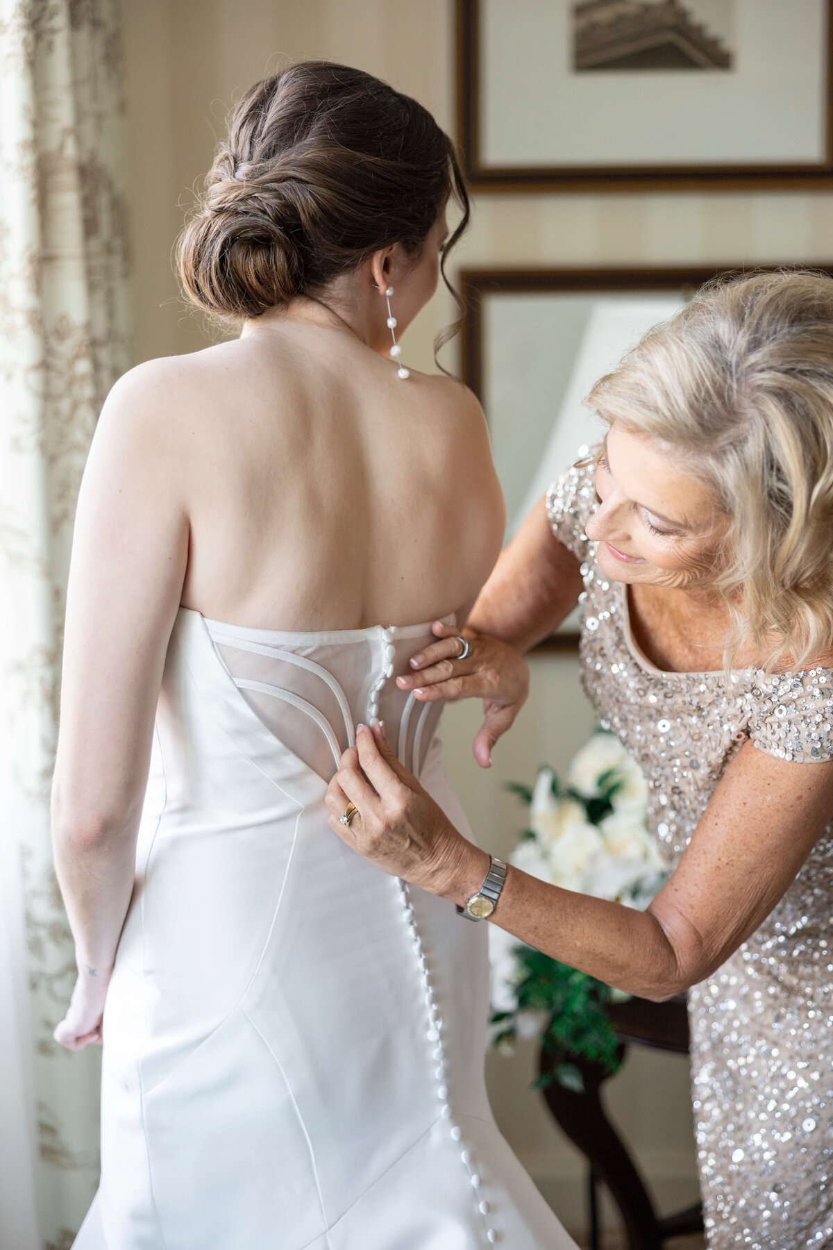 A woman in a sequined dress helps a bride button her strapless white wedding gown. The scene is set indoors with a framed picture on the wall and a floral arrangement in the background.