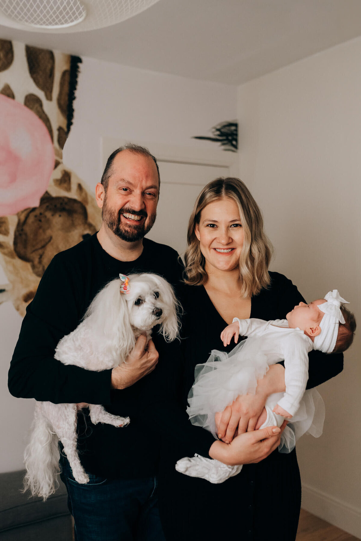 A mother and father hold their newborn daughter and their little white dog in a safari themed nursery