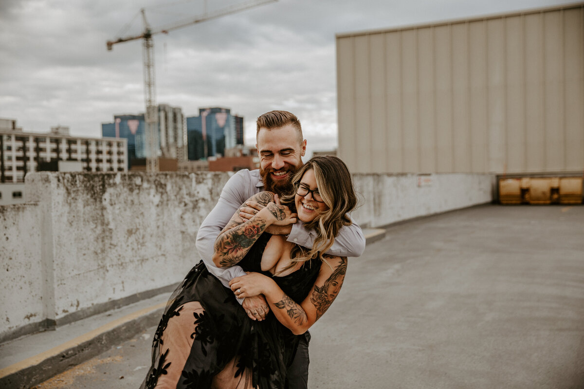 Engagement photoshoot at the top of a London, Ontario parking garage. The man is standing behind the woman, tight in an embrace with lots of movement. They are both smiling in this candid photo.