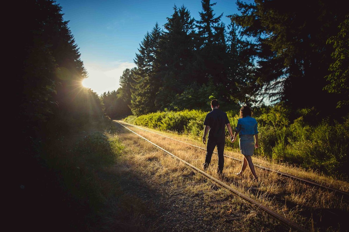 Engaged couple walks on train tracks during sunset