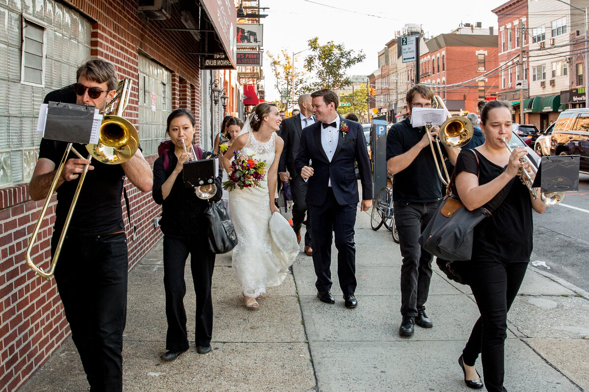 A bride and groom walking down a sidewalk with a band playing around them.