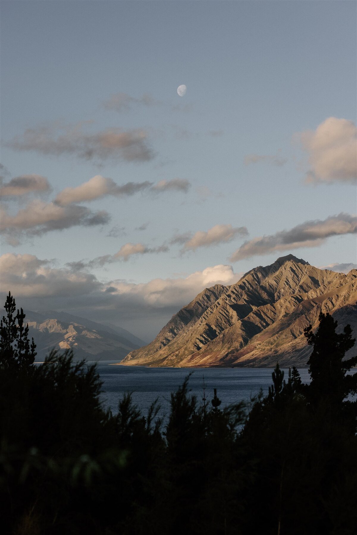 Dramatic landscape of Lake Hāwea at dusk, with moonlight casting a soft glow over rugged mountains and water, framed by treetops.
