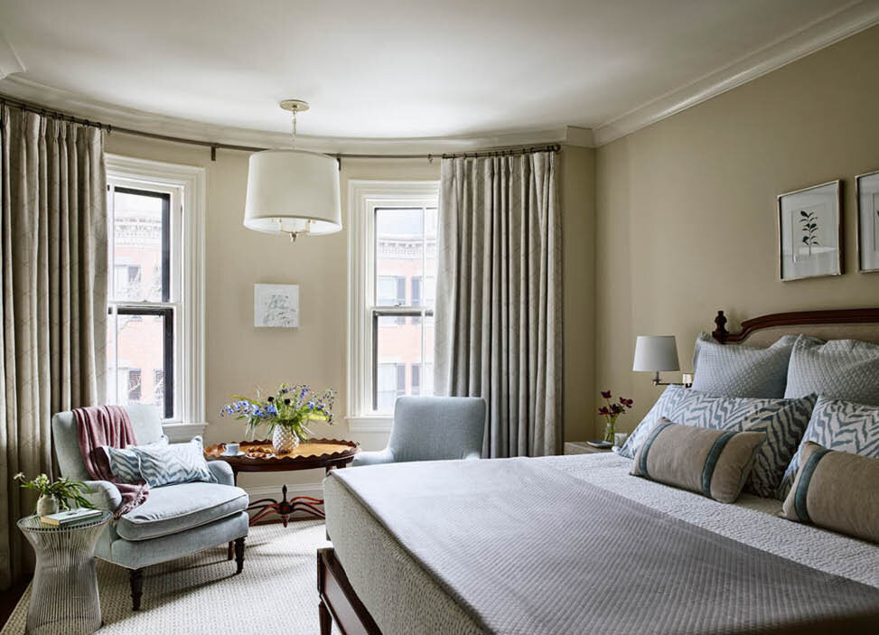 A classic bed and bedding, custom upholstered chairs in a curved nook, and custom drapery with a hanging shade pendant create a timeless bedroom. By Sarah Scales.