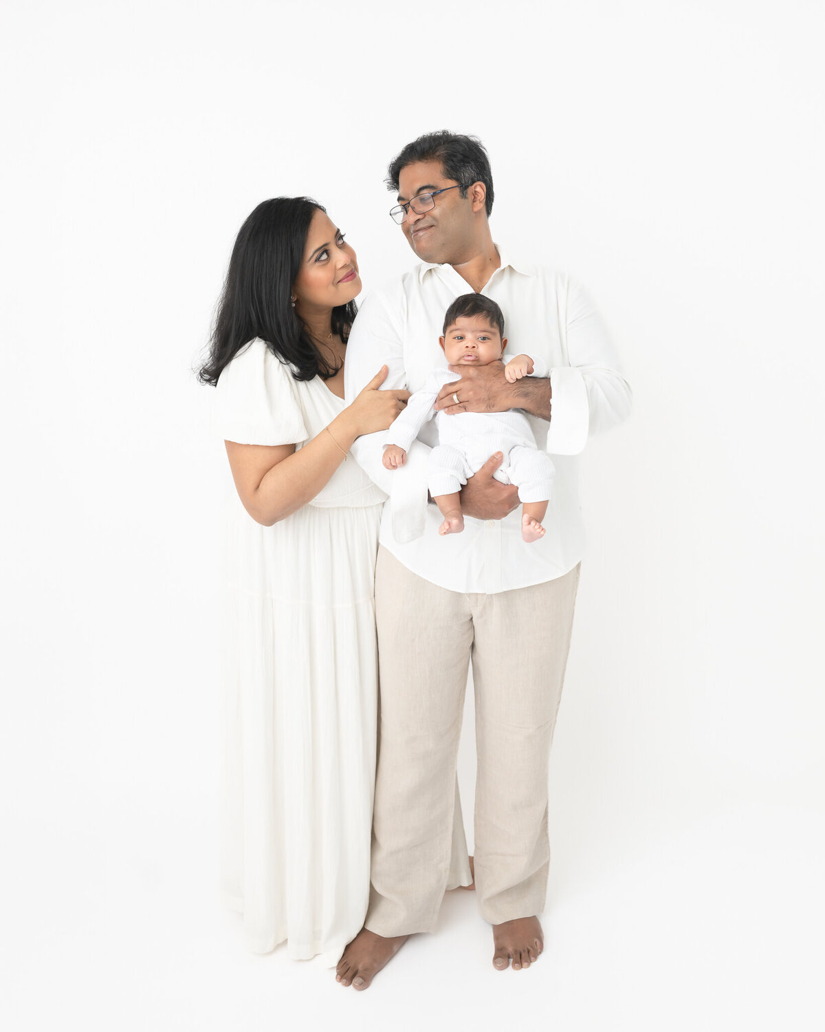 New parents dressed in white with new baby also in white in studio portrait on white backdrop
