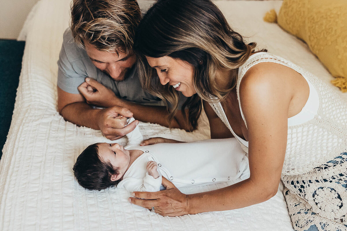 adorable family of three sit together on a bed while mom and dad grin at their beautiful new baby girl with a full head of hair