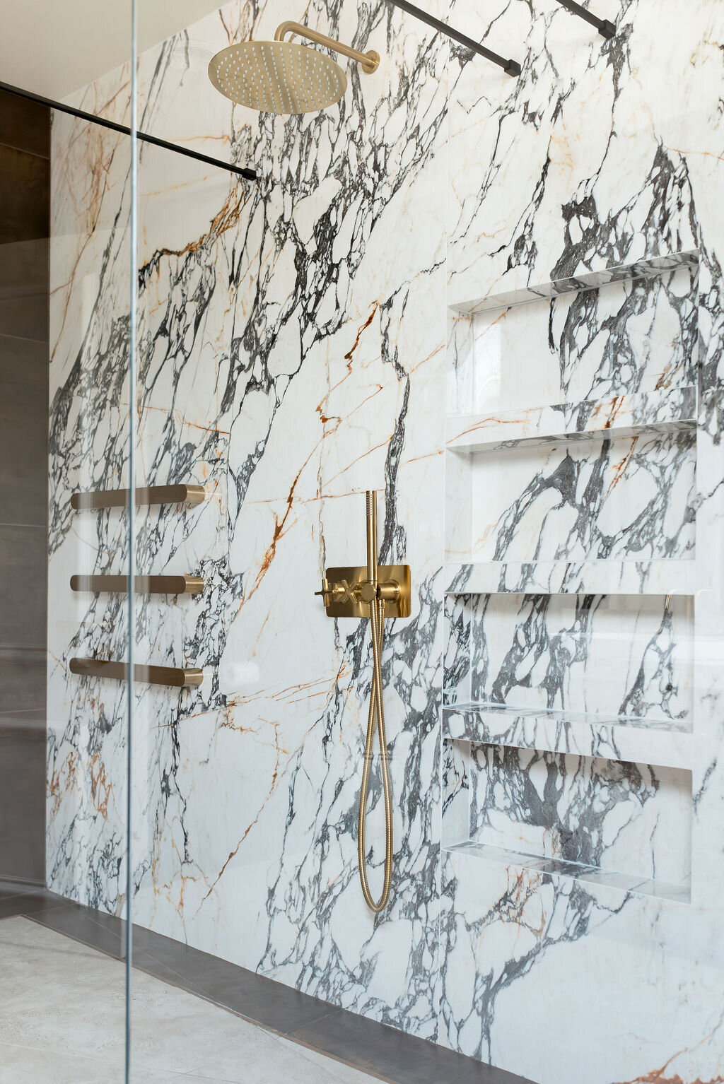 A luxurious shower features a marble wall with black and gold veining. Three gold shelves are mounted on the left side of the wall. A gold rainfall showerhead and a handheld shower attachment are on the right, with modern fixtures and controls.