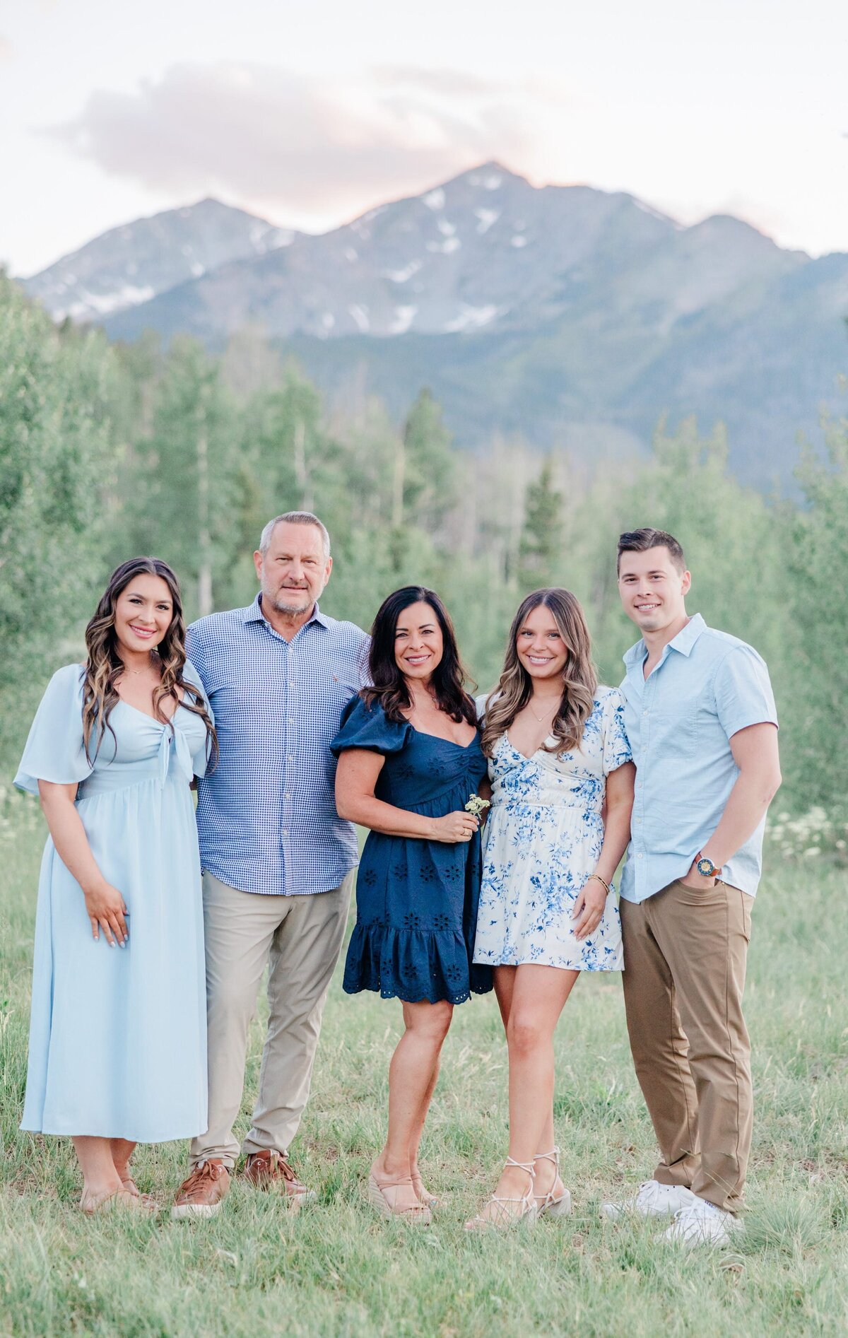 A happy mom and dad stand smiling with their three adult children in a mountain meadow