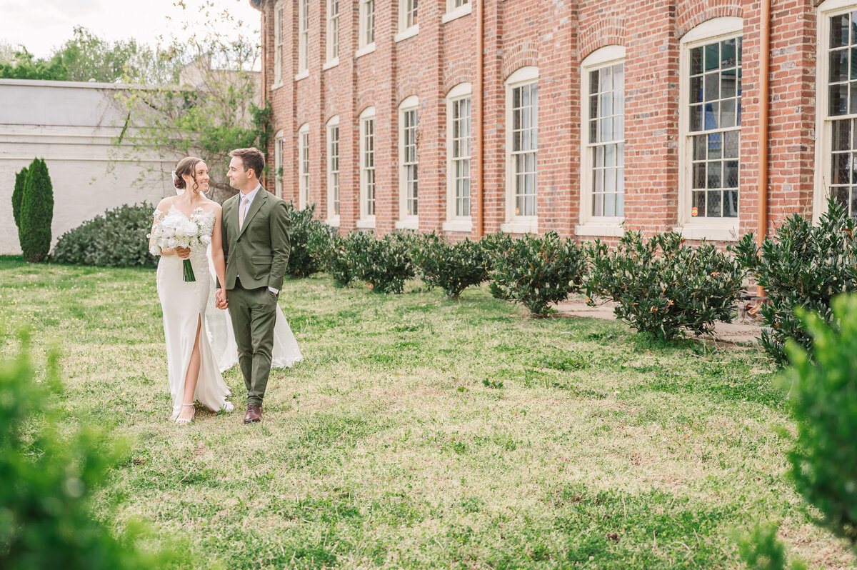 A loving couple on strolling next to their classic brick venue in Virginia during June as they enjoy their Raleigh wedding photography session by JoLynn Photography