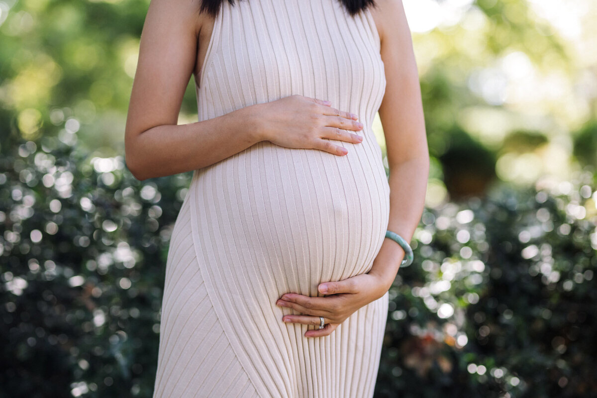 baby bump of pregnant woman wearing white dress and hands on her belly standing in front of greenery at Berkeley Gardens