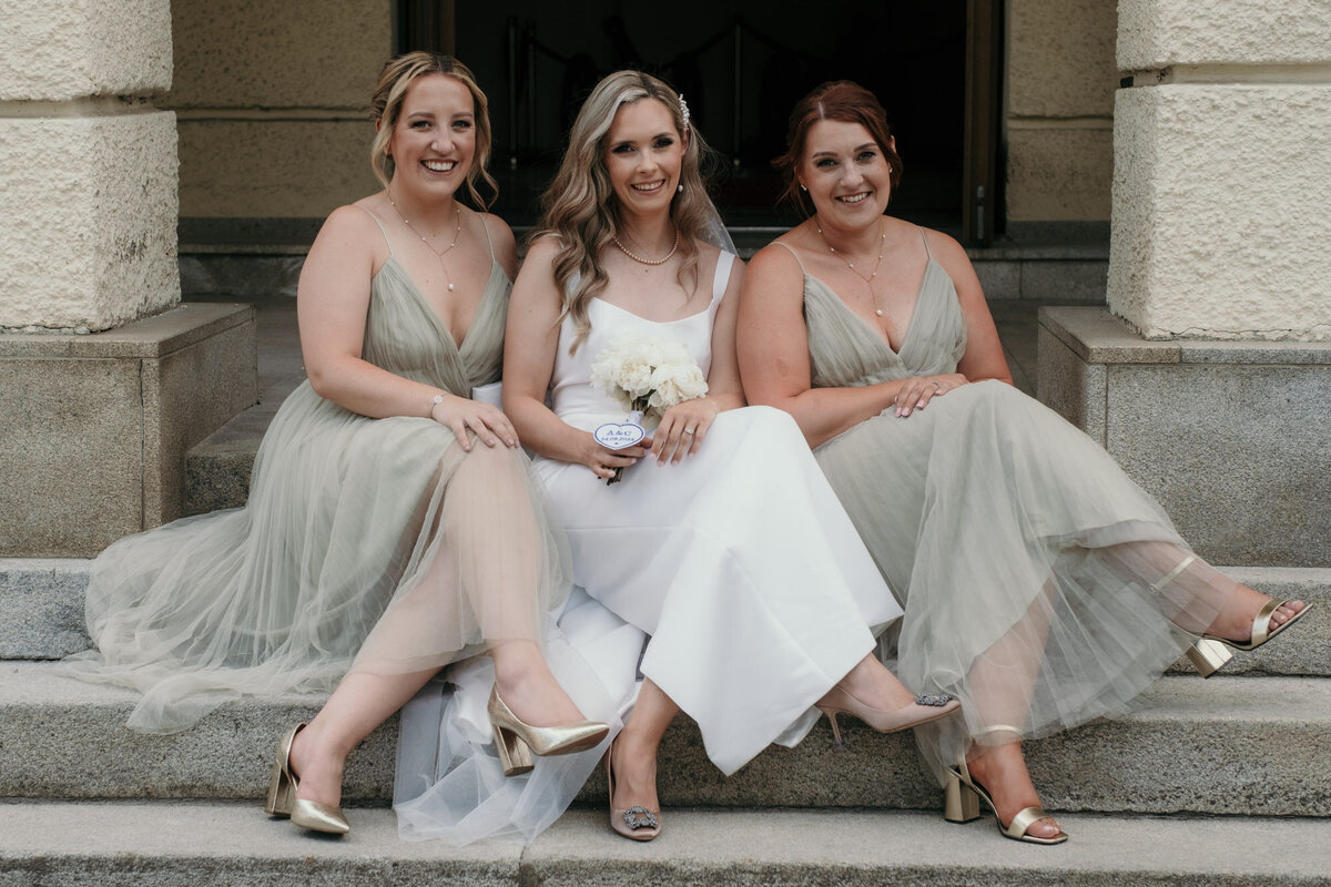 Mit überschlagenen Beinen sitzend posieren die Braut und ihre zwei Brautjungfern auf einer Steintreppe.