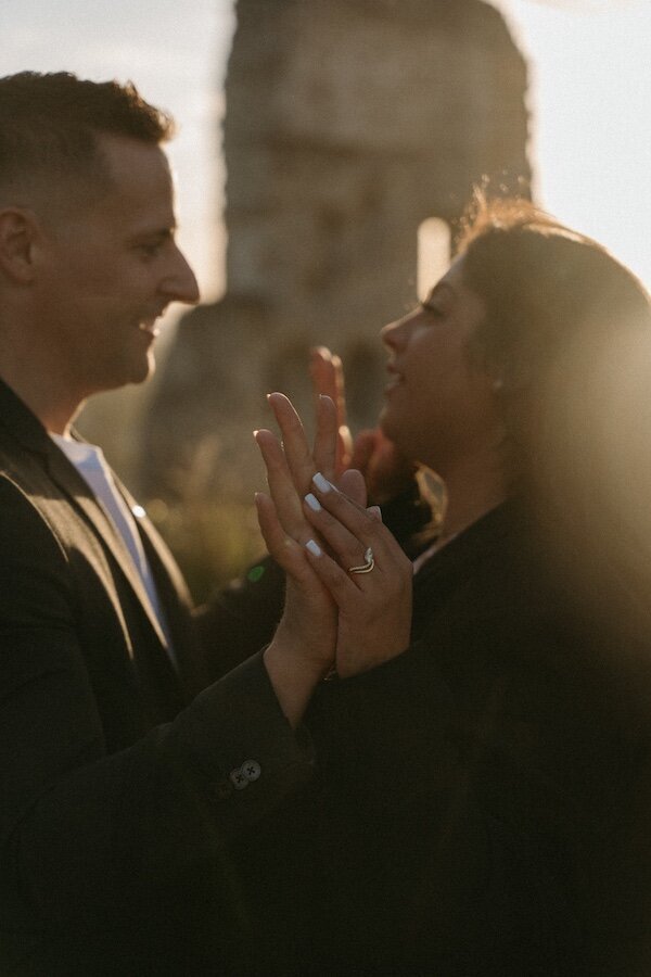 Jeu complice entre Anissa et Brendan, leurs mains entrelacées laissant entrevoir la bague de fiançailles, au coucher du soleil. Séance engagement par Kamescop en Normandie.