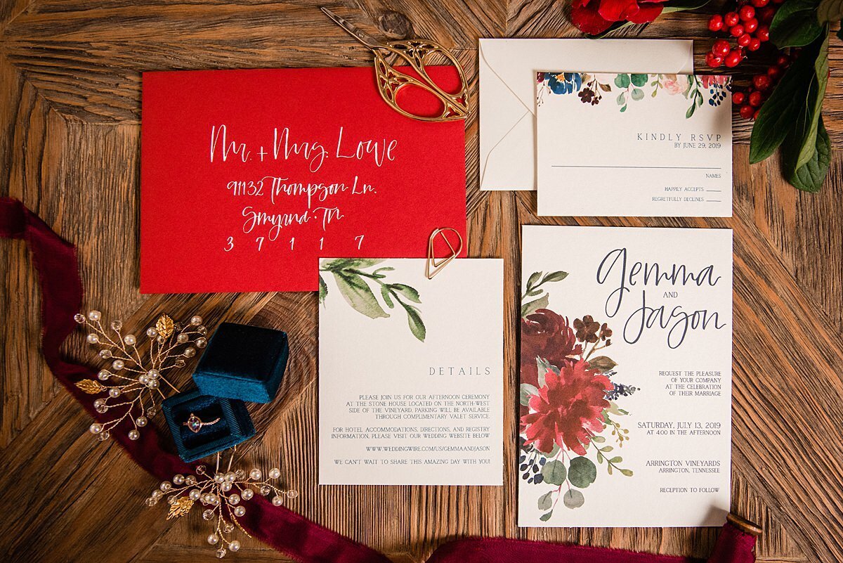 A burgundy and red stationery suite with watercolored flowers and greenery. A bright red envelope with a hand calligraphy written address written in white accented by red hypericum berries a dark teal velvet ring box  with a vintage rose gold sapphire engagement ring and gold and pearl hair jewelry embellishments with a raw silk burgundy ribbon on a farm table at Arrington Vineyards.