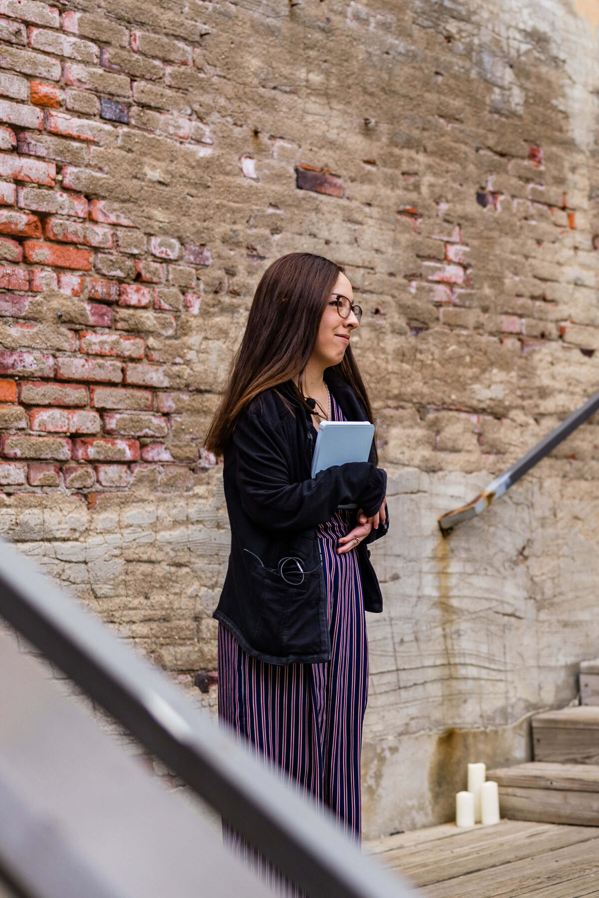 A woman officiant with long brown hair, wearing glasses, a black jacket, and striped dress stands by a weathered brick wall, holding a folder. There are candles at the bottom of nearby steps.