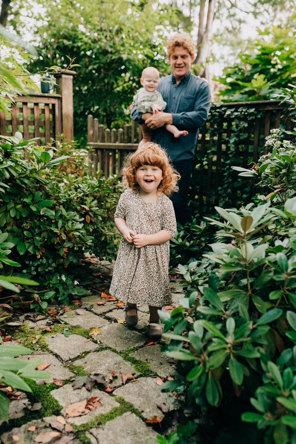 A toddler girl with red hair stands in her back yard in front of her father who is holding her baby sister
