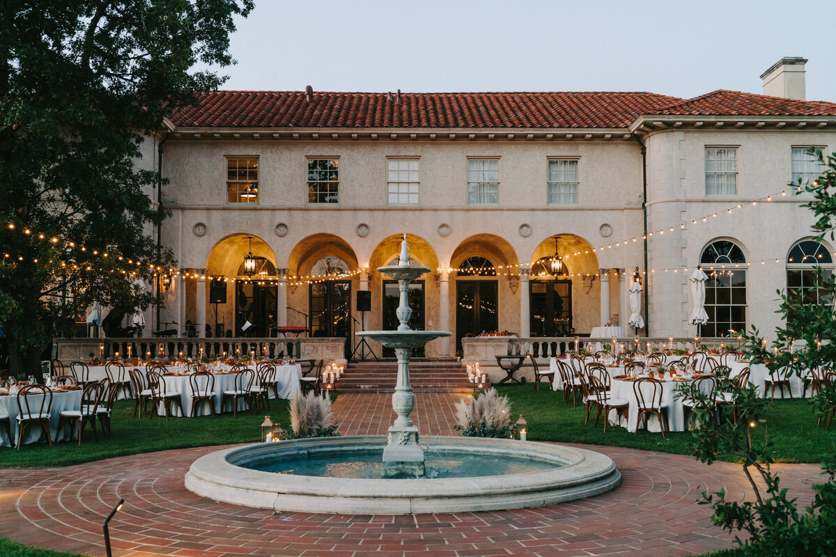 A photograph in color showing the outdoor dinner reception space on Colby and Jack’s wedding day at Commodore Perry Estate in Austin, Texas. The backdrop is a two story, historical mansion with a fountain in the foreground. The round reception tables are spread out on either side. It is just after sunset and the cafe lights overhead are glowing. Wedding photography taken by Stacie McChesney of Vitae Weddings.