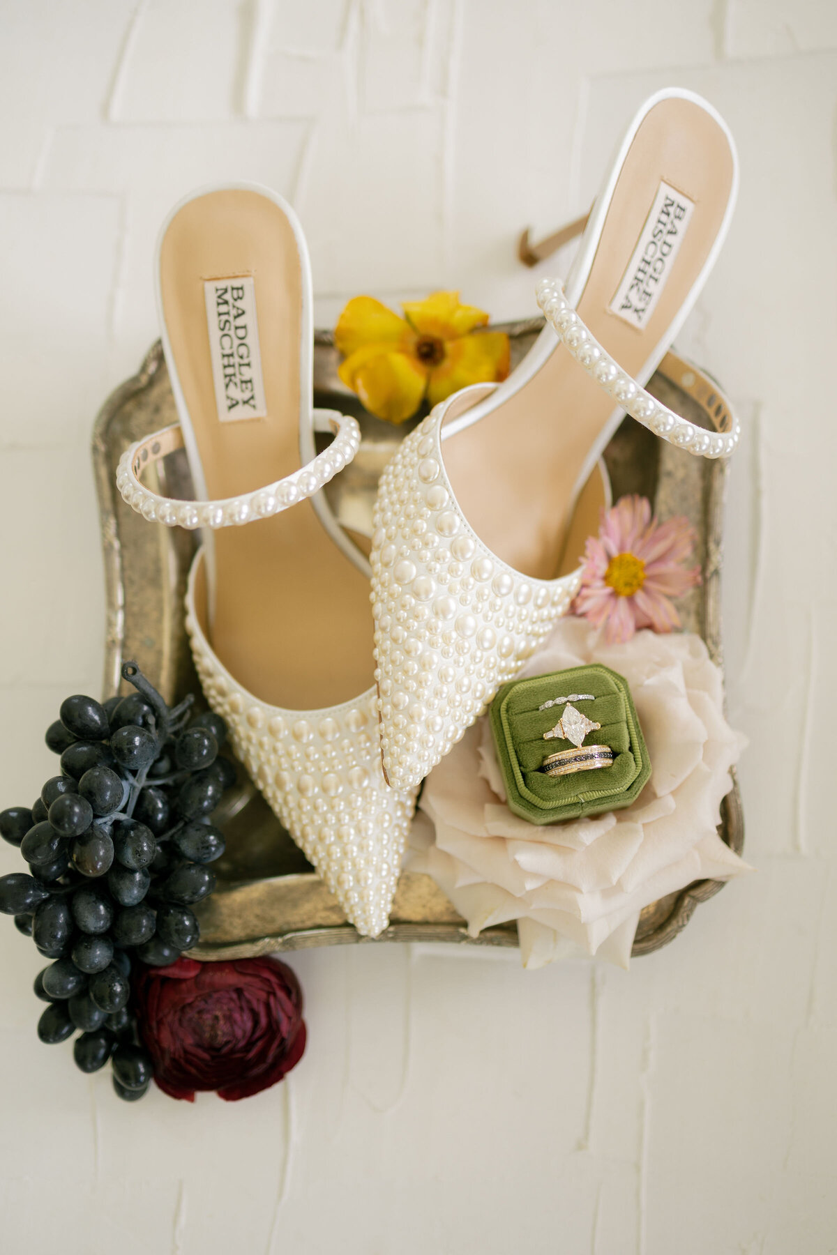 Badgley Mischka pearl-embellished bridal heels styled with a green velvet ring box holding a marquise diamond ring, surrounded by florals and fruit on a vintage silver tray for luxurious wedding flat-lay inspiration.