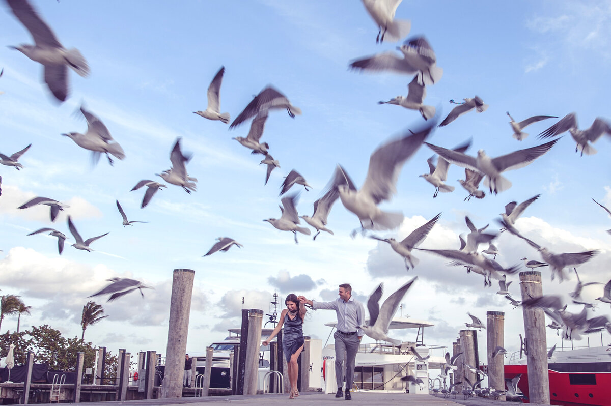 Couple holding hands and walking along a dock in Miami, surrounded by a flock of seagulls taking flight, with boats and a vibrant sky in the background.