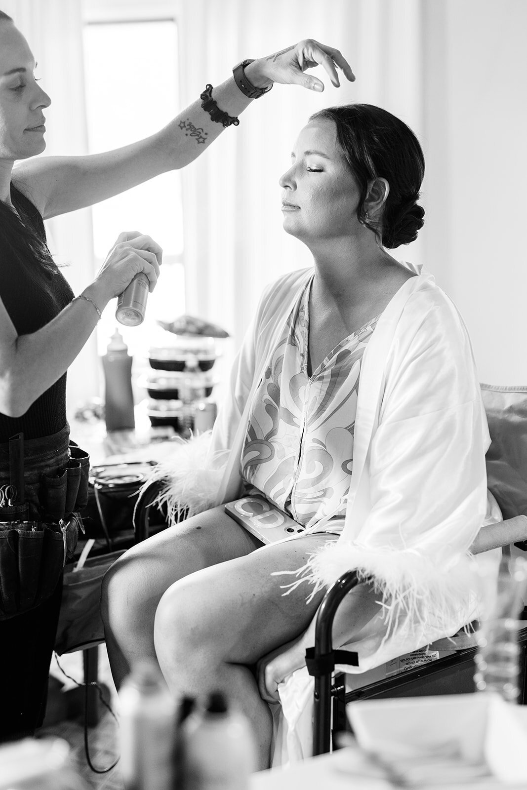 A black and white photo of a bride, dressed in a feathered robe, getting her makeup done. 