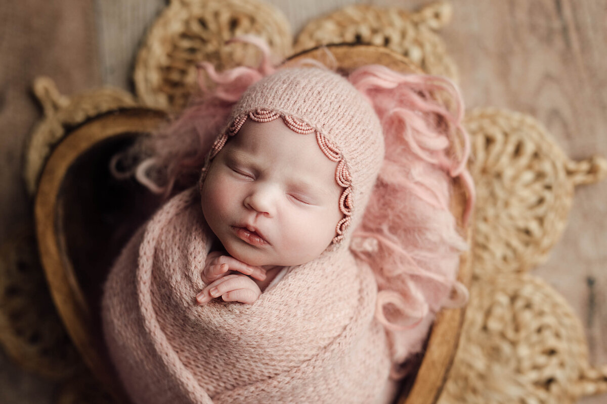 Baby girl sleeping in a wooden heart bowl. She is wrapped in a light pink knit swaddle with matching bonnet with a beaded trim. Her hands are folded under her chin and peeking out of the swaddle.