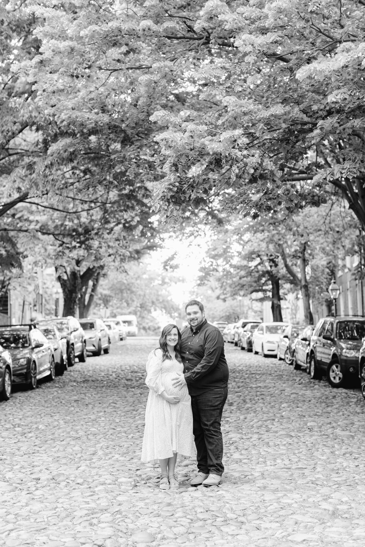 A Northern Virginia Maternity Photographer photo of a pregnant couple smiling at the camera on cobblestone streets in Old Town Alexandria VA