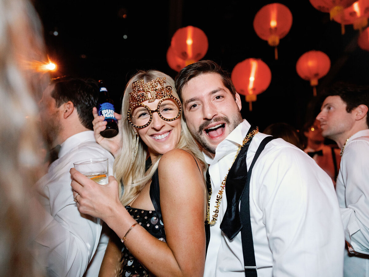 A happy couple poses for the camera, dressed in festive attire with drinks in hand, enjoying the wedding celebration.