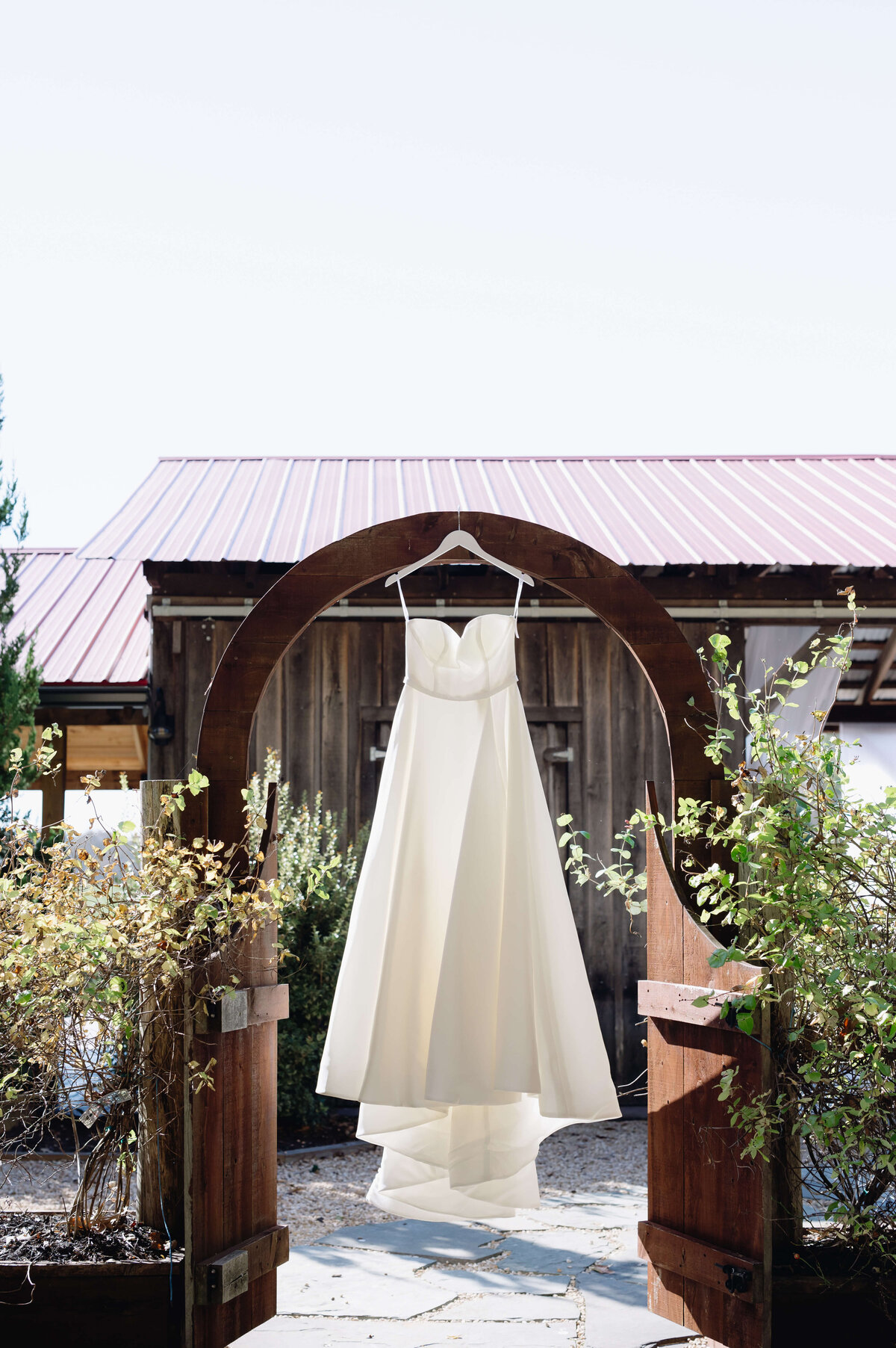 satin wedding dress hanging in a garden on a walkway arch with a cute shed behind it