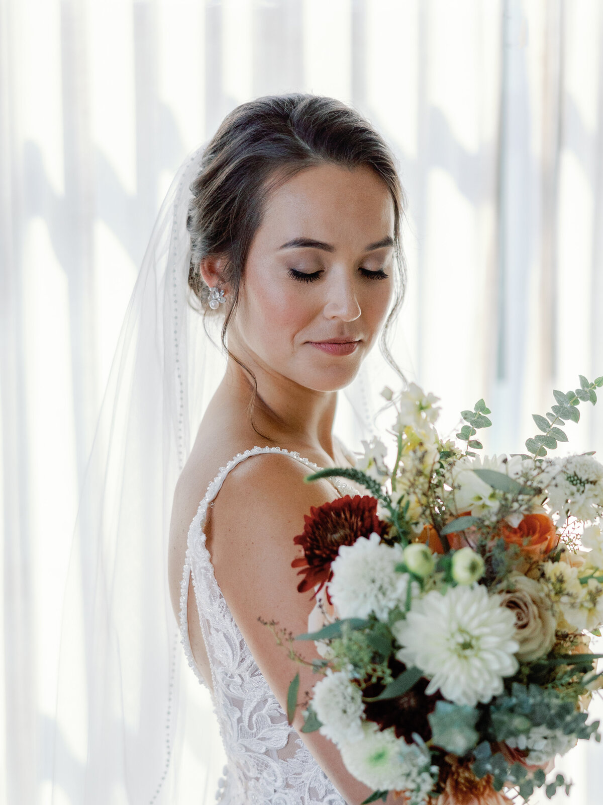 A bridal portrait of a bride holding and looking down at her bouquet full of white, red, and orange flowers.