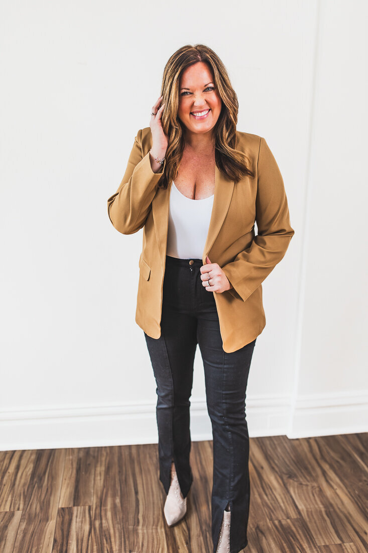 A professional headshot or website image of a smiling woman with long wavy brown hair wearing a camel-colored blazer over a white top and dark pants.