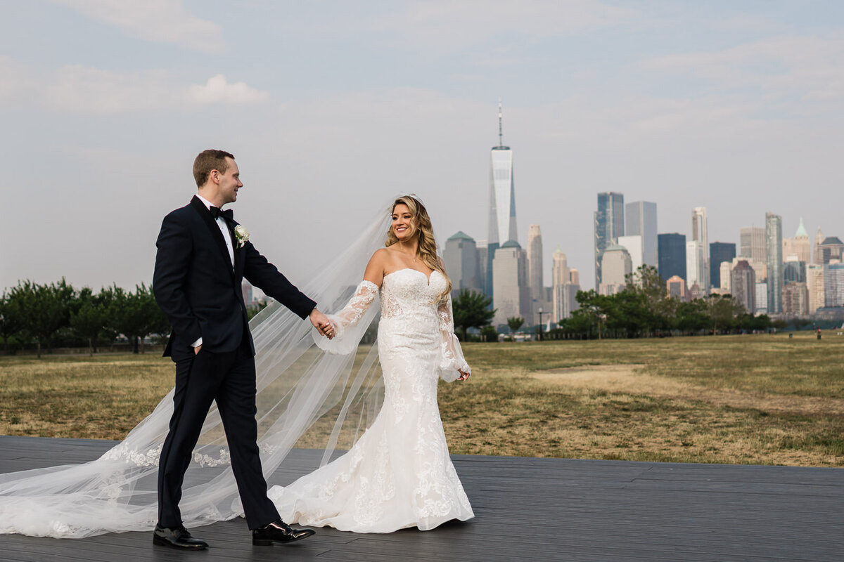 Bride and groom walking together outdoors at Liberty State Park, with the bride’s veil flowing and the New York City skyline in the background.
