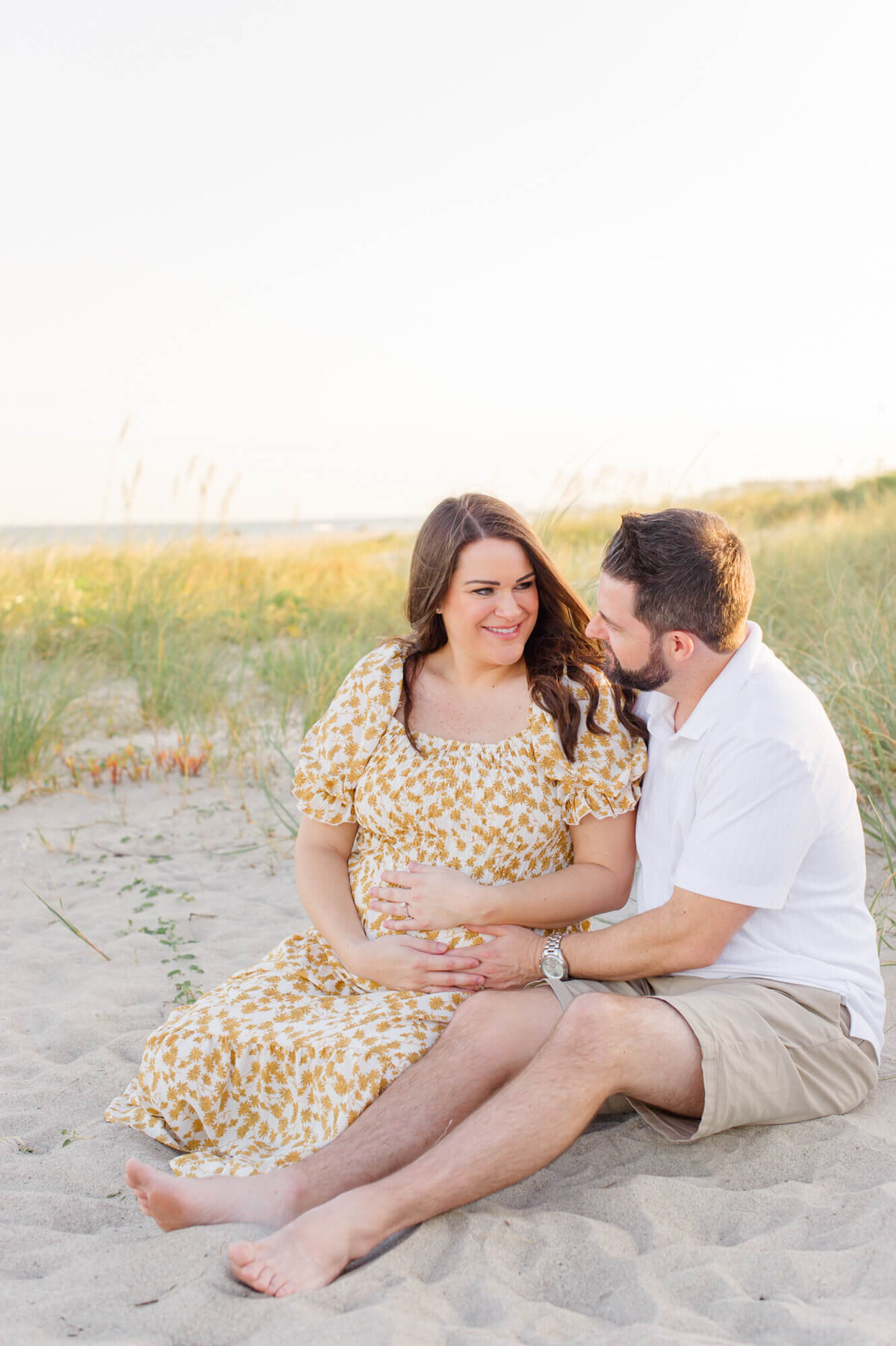 Expectant parents sit in the dunes at sunset holding mothers pregnant belly at golden hour