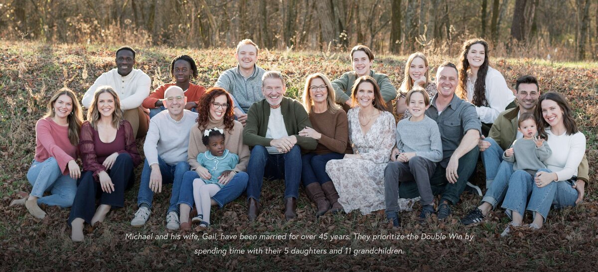 Large multigenerational family sitting together outdoors in a wooded area, arranged in two rows and smiling at the camera.