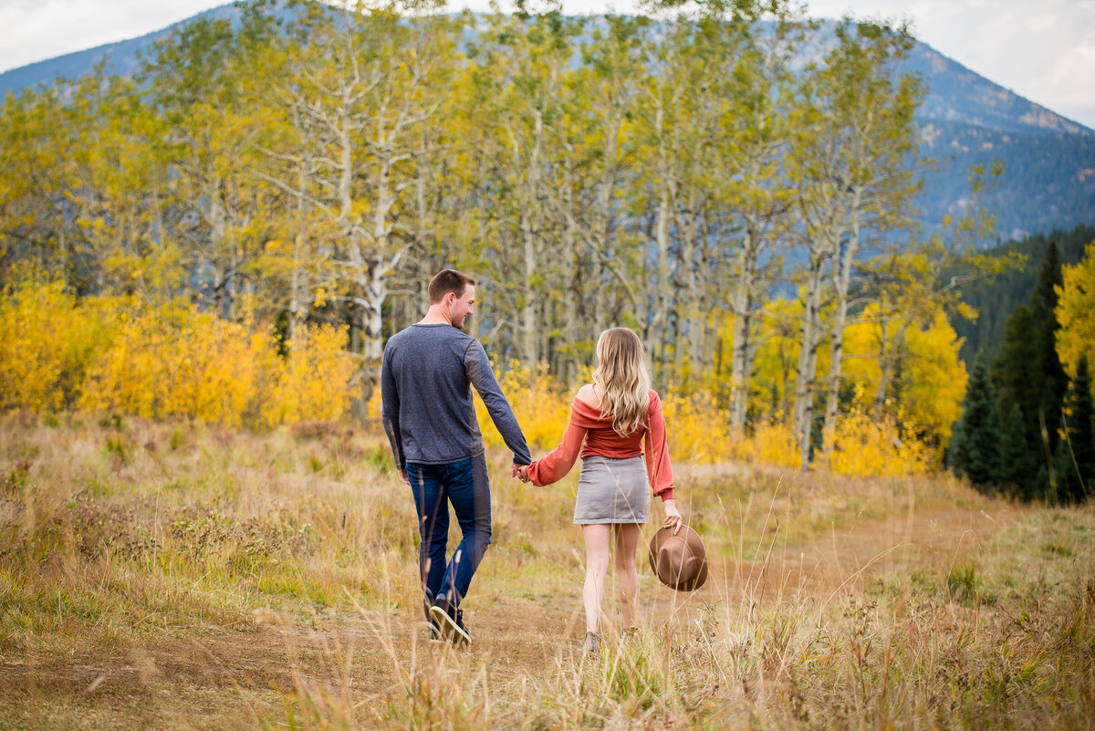 A man and woman walk away from the camera holding hands in a fall setting. The woman holds a hat.