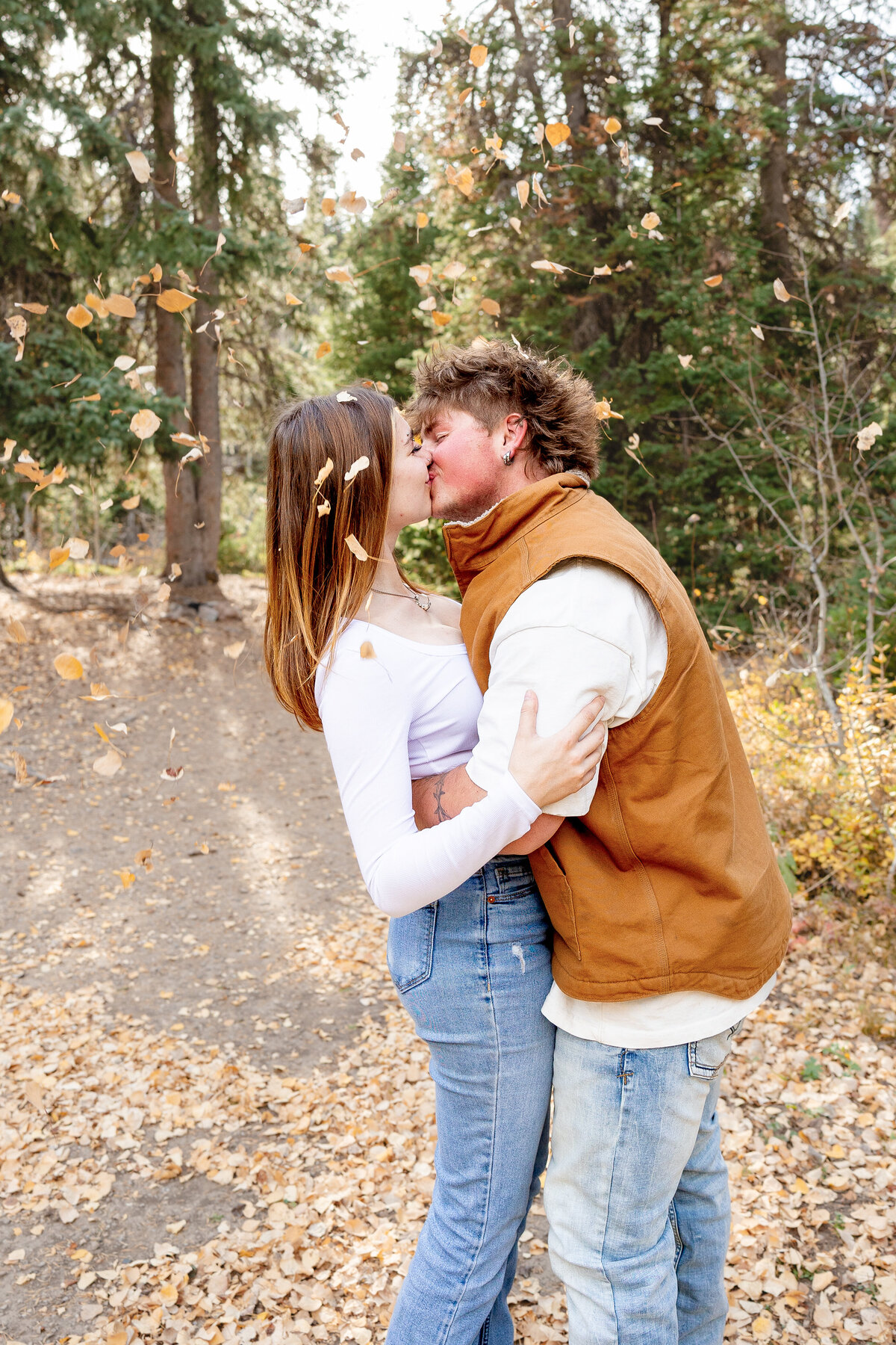 A couple kisses in the middle of a forest trail as golden autumn leaves fall around them, capturing a playful and romantic fall moment.