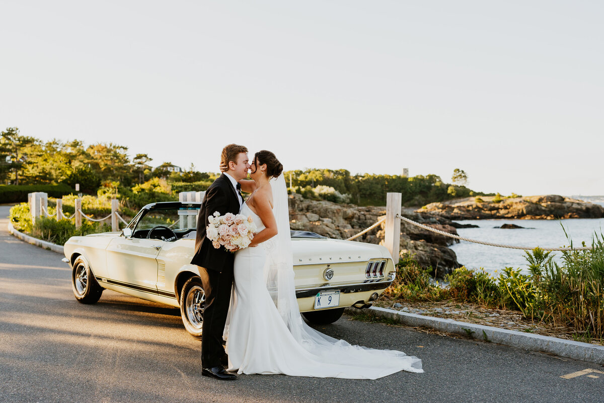 bride and groom embracing with old white car and ocean in the background