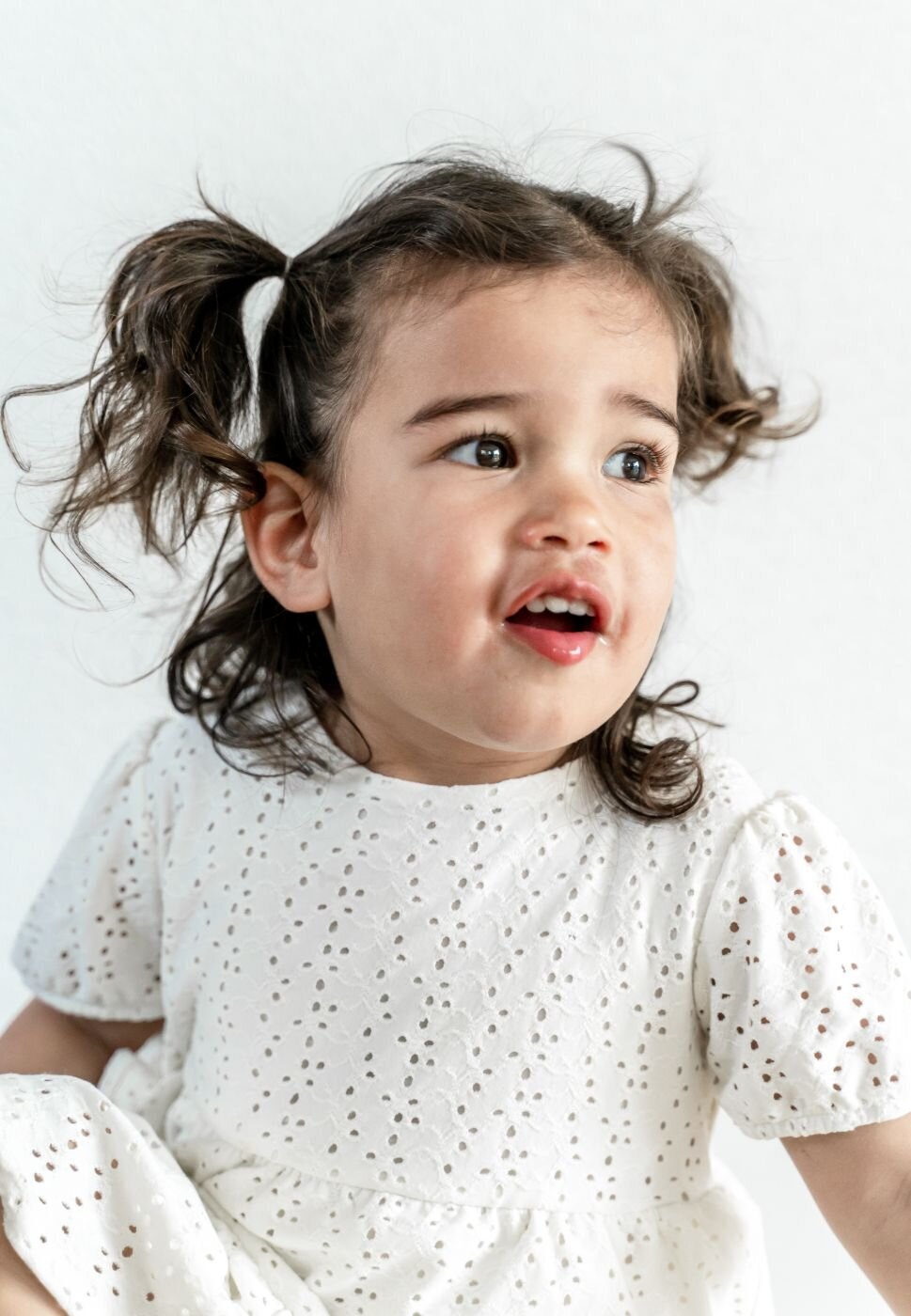  A close-up color portrait of a little girl with a surprised and happy expression. Her curly brown hair is styled in pigtails, and she is looking off to the side with her mouth slightly open. She is wearing a white dress with an eyelet pattern and is standing against a simple white background.