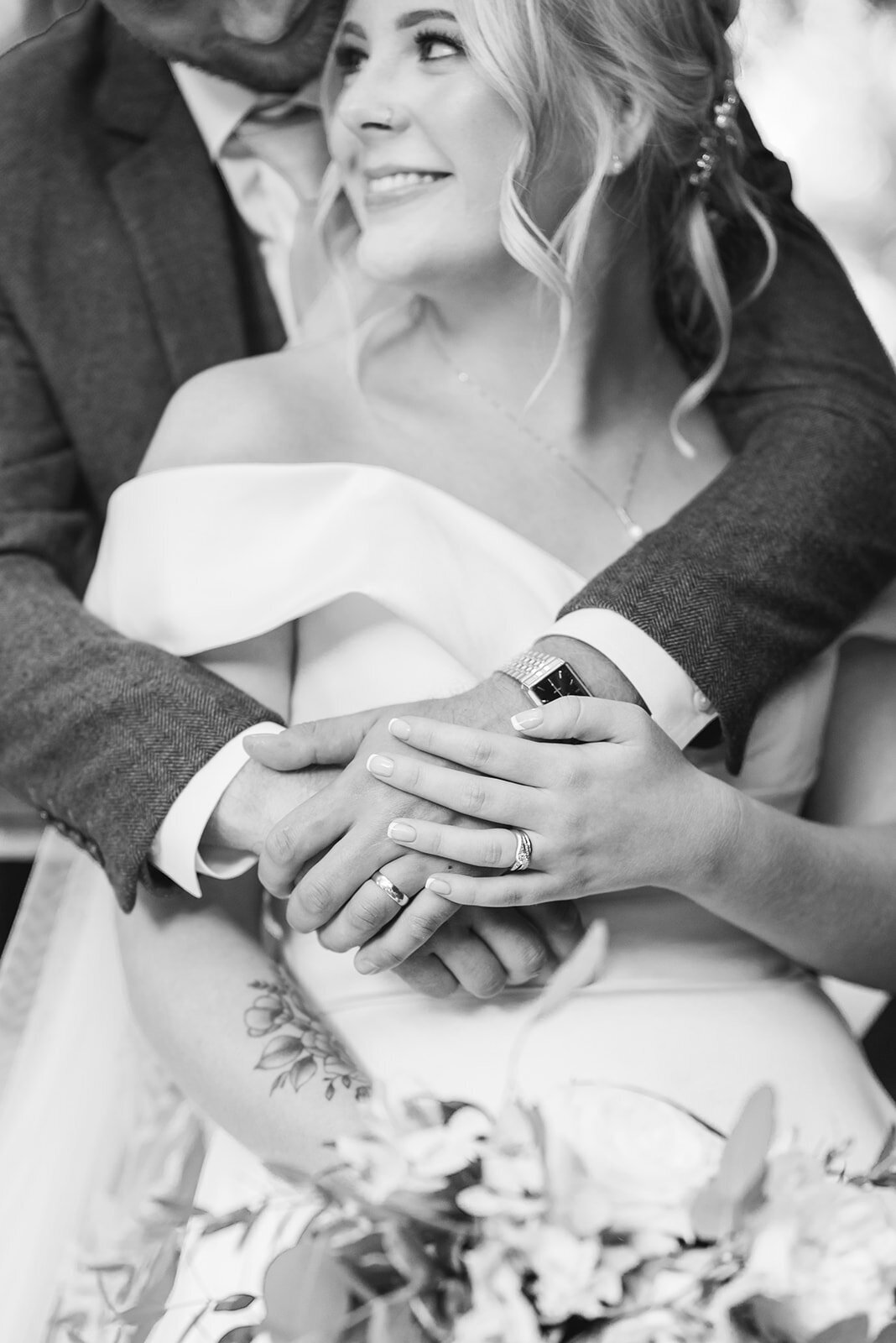 Black and white image of a bride and grooms hands, focusing on their rings. The groom is behind the bride and has his arms around the brides shoulders. The brides hand is placed gently on top of the grooms hand.
