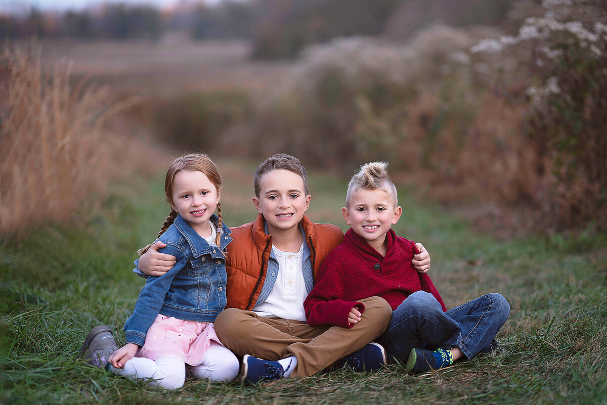 Siblings sitting in a field for family photos. Big brother is in the middle and has his arms wrapped around his little brother and sister. Captured by Liza Nicole Photography, Philadelphia and Main Line family photographer.