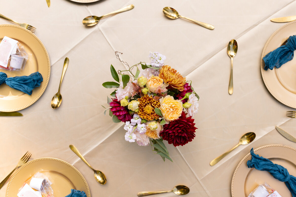 Wedding reception table setting with flowers in the middle of a round table , and spoons and plates surrounding it.